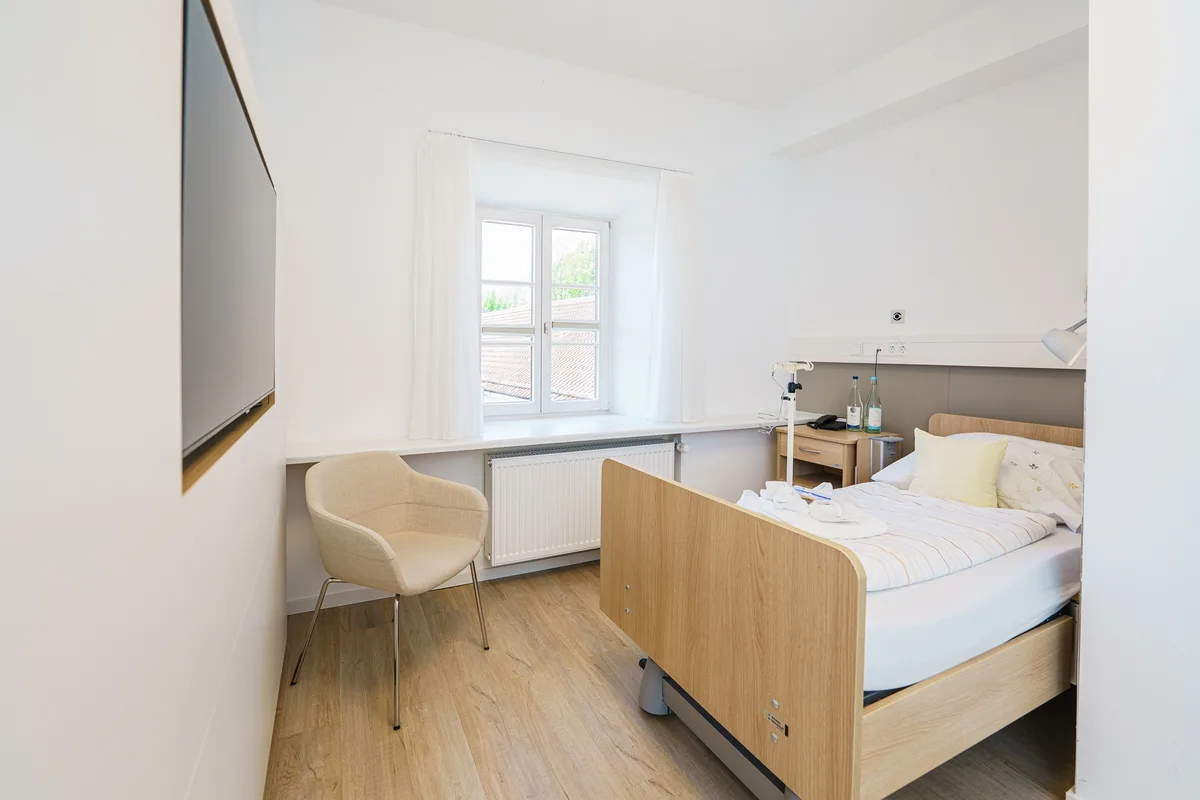 A patient room with a wooden-framed hospital bed, an armchair and a bright window.
